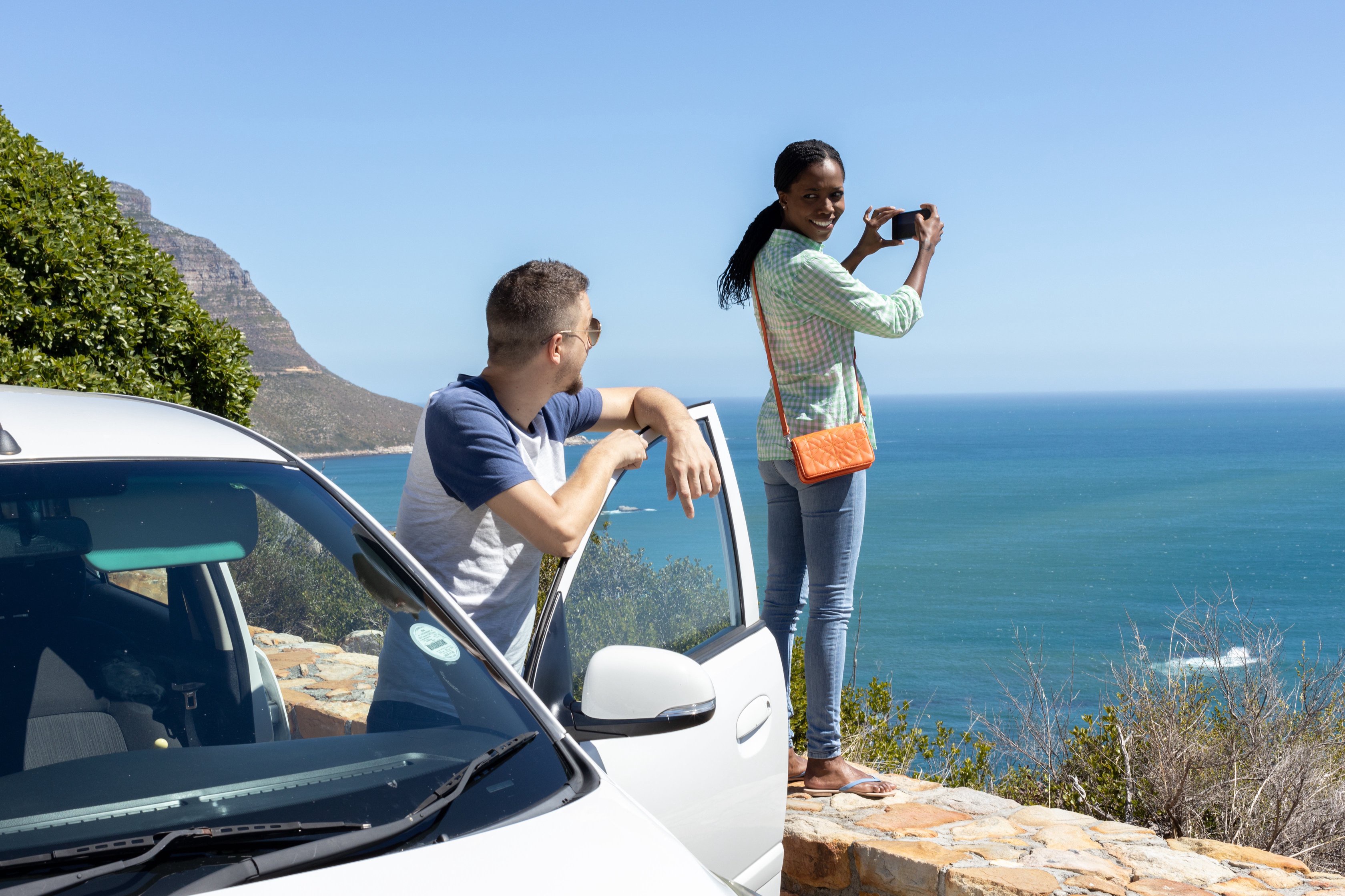 A person stands near a car taking a photo of a scenic ocean view, another person leans on the car door, enjoying the view