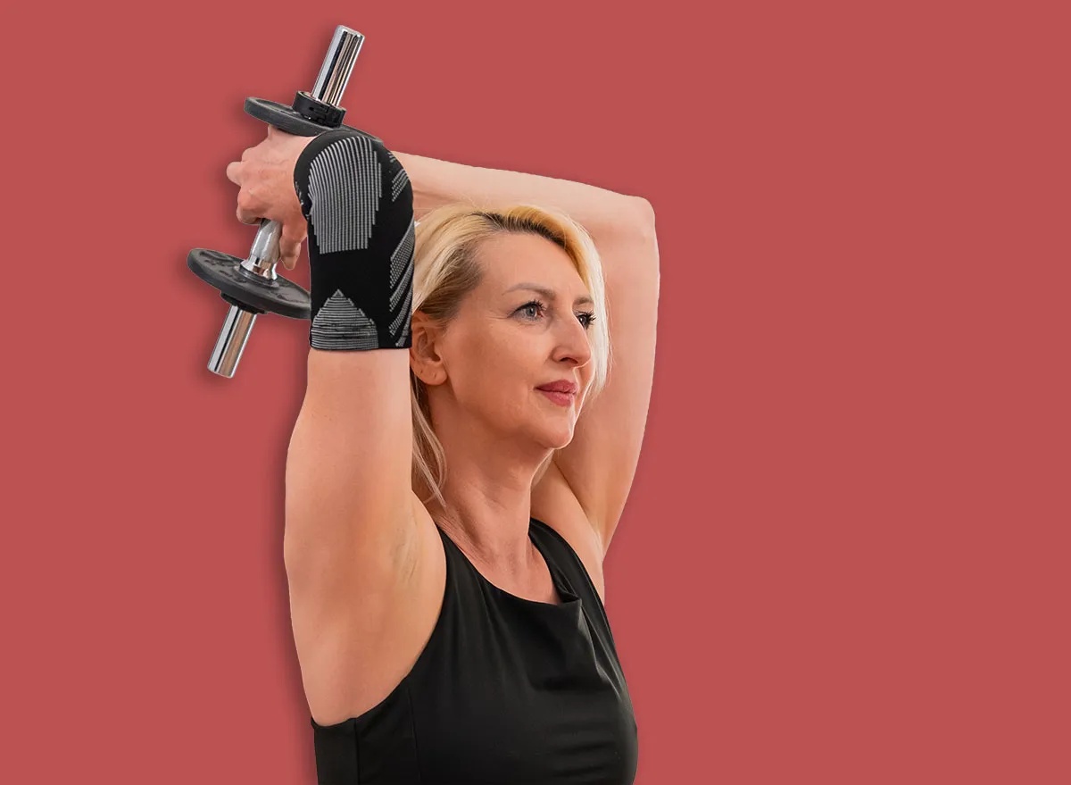 Focused blonde middle aged woman sitting on floor mat lifting dumbbell behind head to train triceps during home workout in modern living room