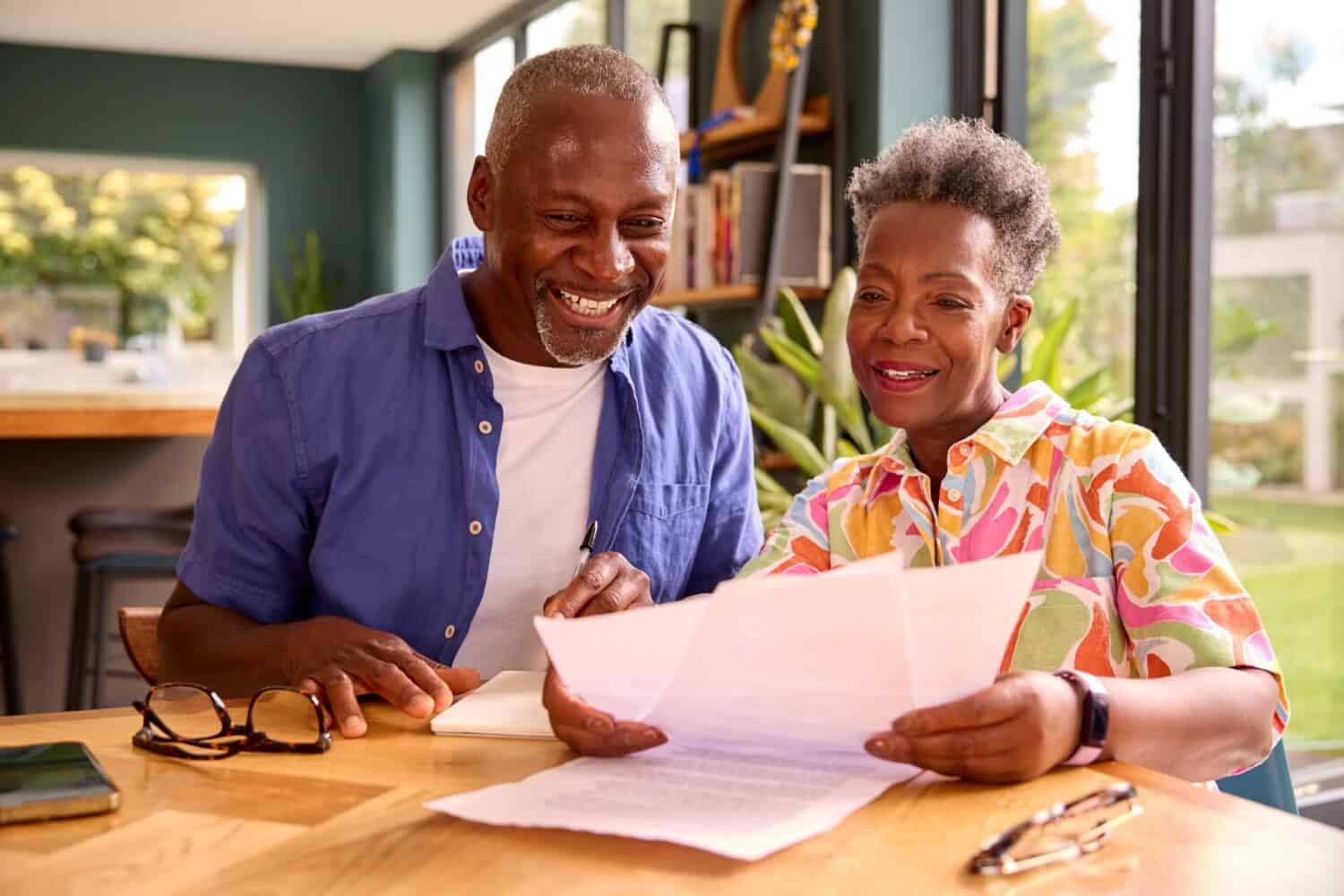A smiling Black man in a blue shirt and a Black woman in a colorful patterned shirt sit at a light wooden table, looking at papers. The man holds a pen. Eyeglasses and a phone are on the table. The background shows a bright, modern interior with large windows looking out to greenery.