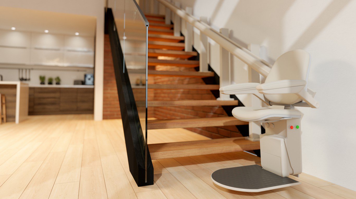 A contemporary home interior showcases a light wooden staircase equipped with a beige, cream-colored stairlift in the foreground, featuring a black footrest and red and green indicator lights. The stairs have a modern glass and dark metal railing on the left and a traditional white wooden railing on the right. In the blurred background, a modern kitchen with light cabinetry and wooden accents is visible.