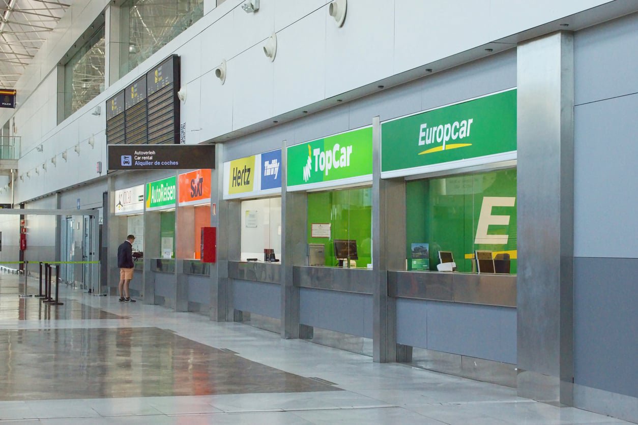 A row of car rental service desks at an airport, featuring company signs for Europcar, TopCar, Hertz, Sixt, and AutoReisen.