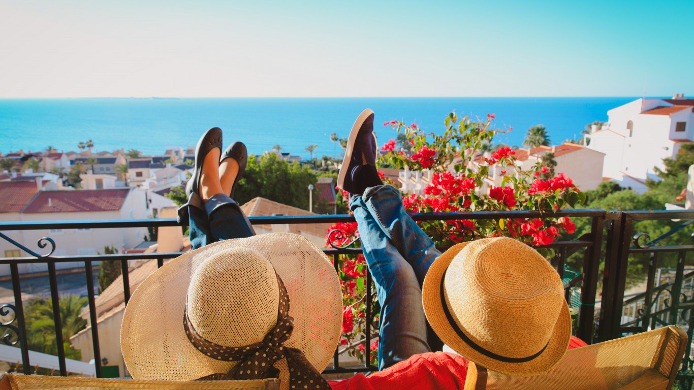 An overhead view shows two people relaxing on a sun-drenched balcony, their feet propped up on a black metal railing. The person on the left wears a wide-brimmed straw hat with a polka dot band, dark casual shoes, and jeans. The person on the right wears a straw fedora, blue jeans, and light-colored shoes, with a red shirt visible. In the background, there's a bright blue sea, a picturesque coastal town with white and terracotta-roofed buildings, and vibrant red bougainvillea flowers.