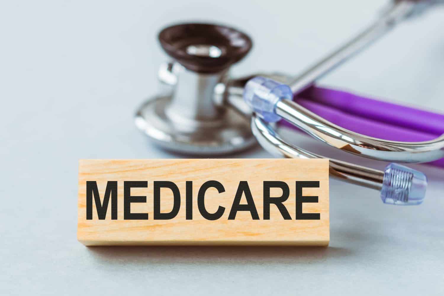 A light brown wooden rectangular block with the word 'MEDICARE' printed in black capital letters. Behind it, a silver and purple stethoscope is partially visible on a light grey surface.