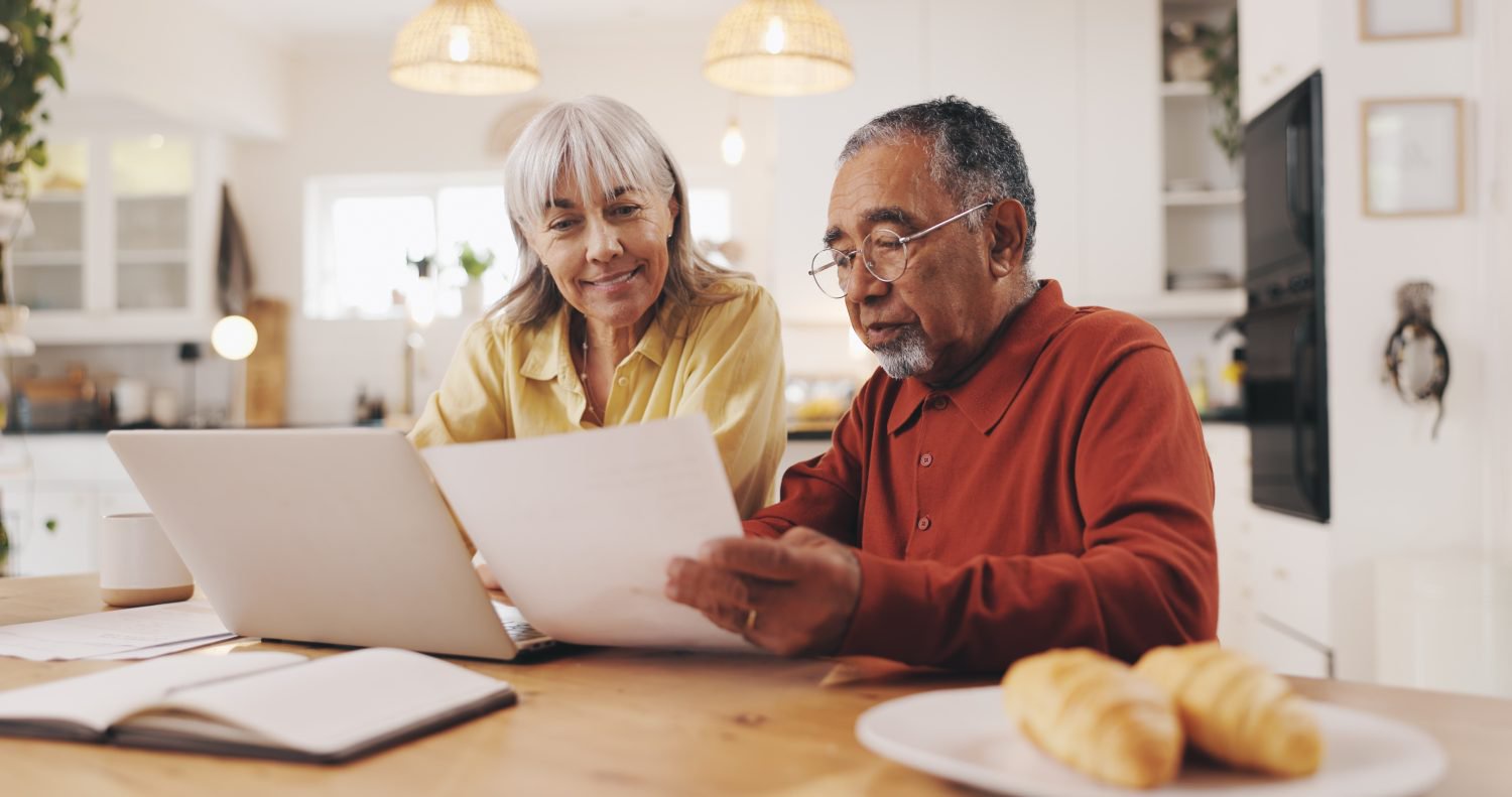 A smiling elderly woman with grey hair and a yellow shirt sits next to an older man with grey hair, glasses, and a reddish-brown polo shirt. They are seated at a light wooden table, focusing on papers held by the man and an open silver laptop. A notebook, a white mug, and a plate with two croissants are also on the table. The background shows a bright, modern kitchen with overhead lighting.