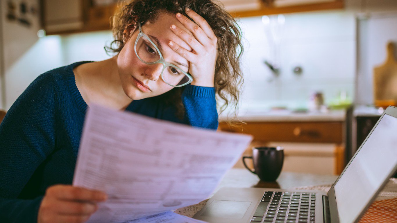 A young woman with curly brown hair and light green framed glasses wears a dark blue sweater, sitting at a table. She holds several papers in her left hand and rests her right hand on her forehead, looking down at the documents with an expression of worry. A silver laptop and a black mug are visible on the table, with a blurred modern kitchen in the background.