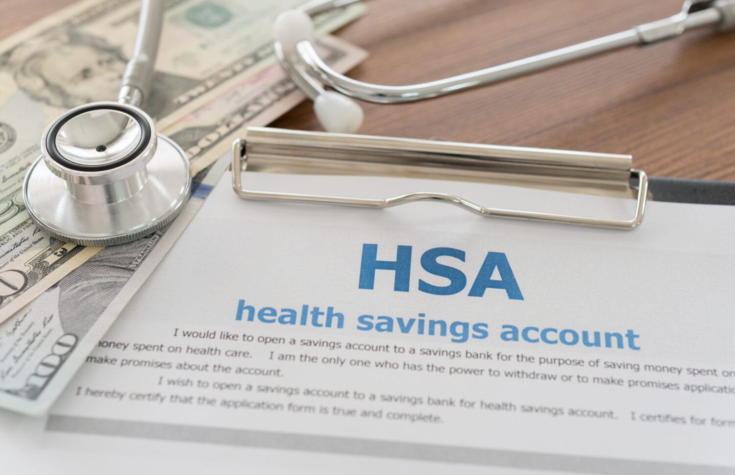 A stethoscope and US dollar bills are arranged on a wooden surface next to a clipboard holding a document. The document prominently displays 'HSA' and 'health savings account' in blue text, with additional fine print visible below.