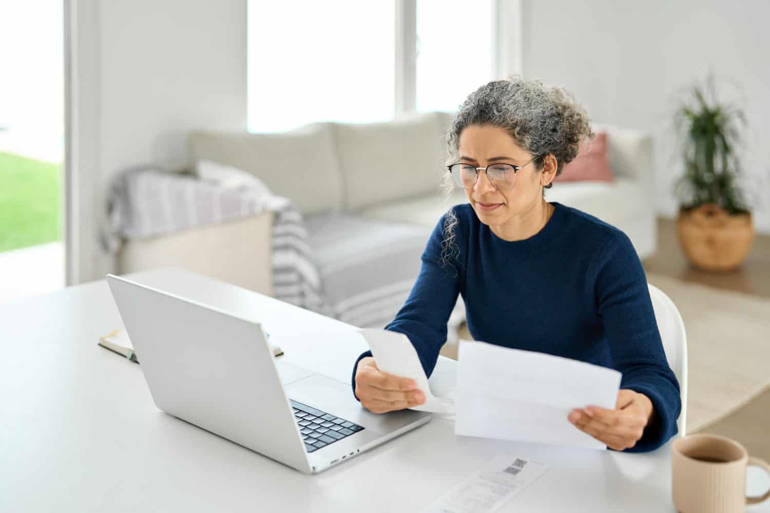 A woman with grey curly hair and glasses, wearing a dark blue sweater, sits at a white desk. She holds white papers in both hands, appearing to review them, with a silver laptop open in front of her. A light beige coffee mug is visible on the desk to her right, and a blurred bright living room with a sofa and a potted plant is in the background.