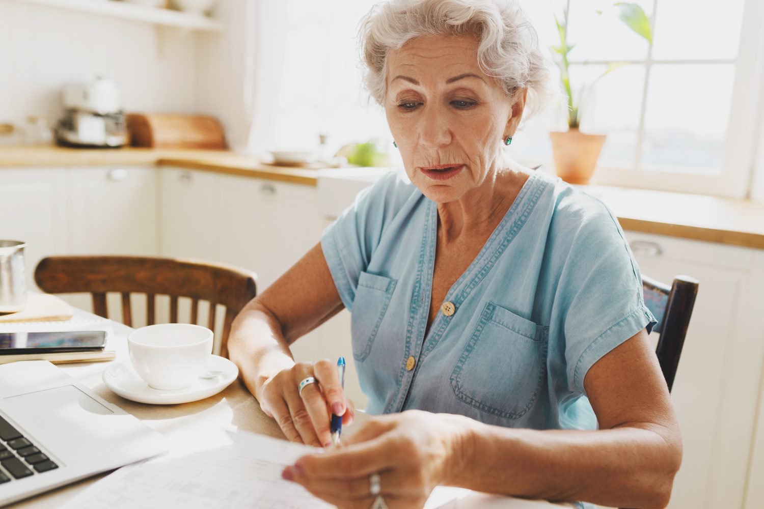 A focused older woman with gray hair sits at a light-colored table in a bright kitchen, intently reviewing papers. She holds a blue pen in her right hand, with a laptop, a white coffee cup, and more documents beside her. She wears a light blue button-down shirt and turquoise earrings. The background shows white kitchen cabinets and a sunlit window.
