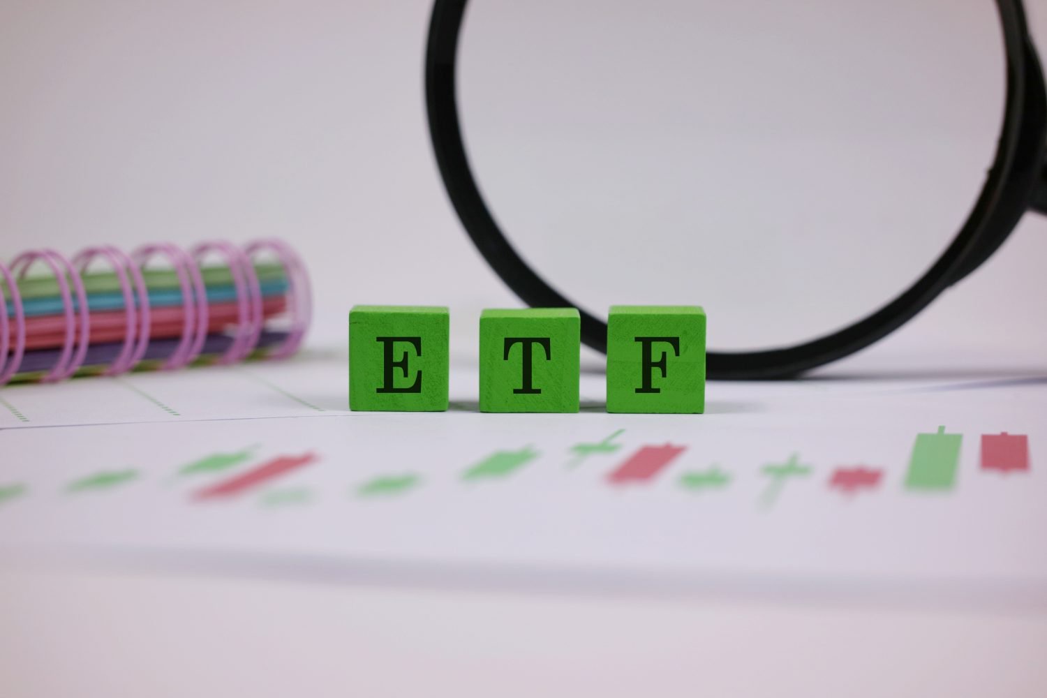 Three green wooden blocks spelling 'ETF' in black capital letters are prominently displayed on a white surface, with blurred red and green financial chart patterns underneath. A blurry black magnifying glass and a colorful spiral-bound notebook with pink, green, and blue rings are in the soft-focus background.