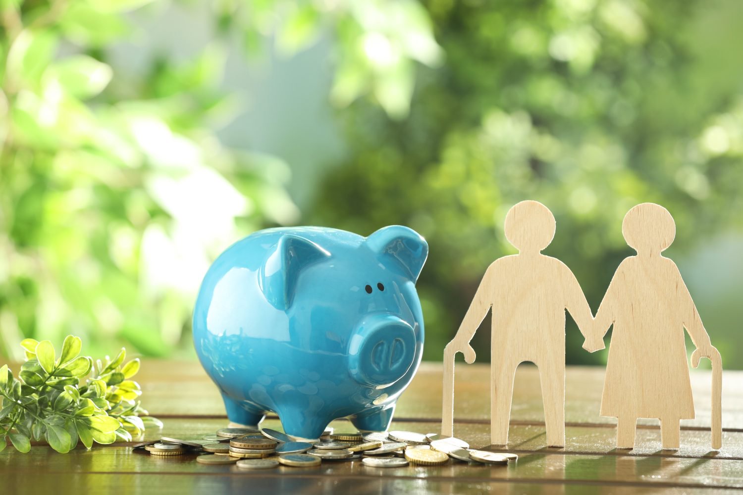 A light blue ceramic piggy bank sits on a scattered pile of coins on a dark wooden surface. To its right are two light brown wooden cutouts of an elderly couple holding hands, both with walking sticks. The background is softly blurred green foliage.