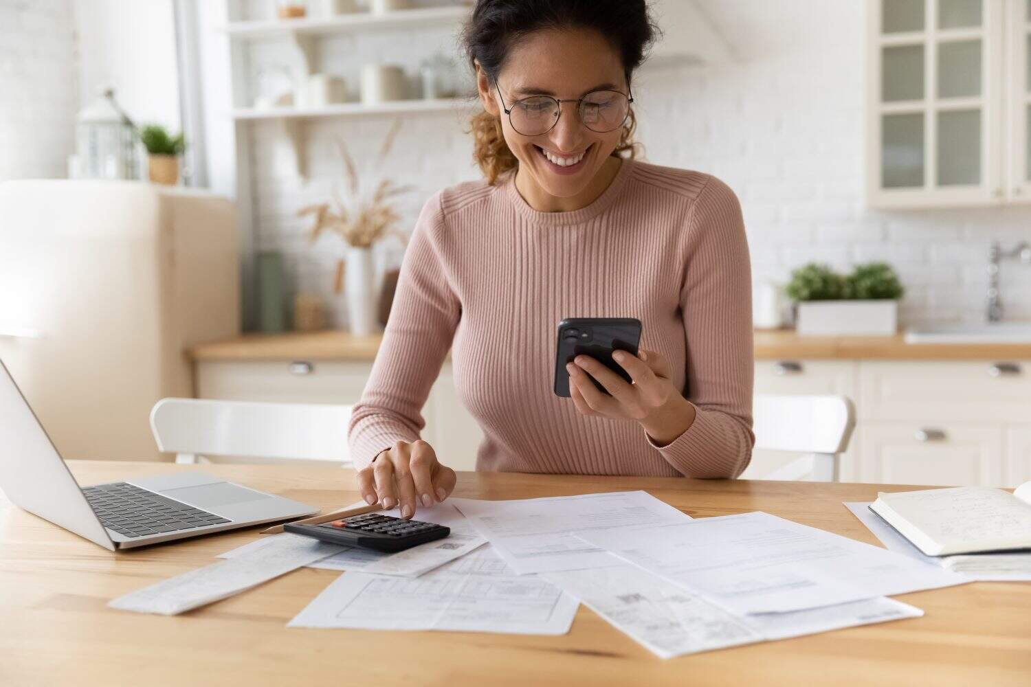 A smiling woman with dark curly hair and glasses, wearing a light pink ribbed sweater, sits at a wooden table. She holds a black smartphone in her left hand and operates a black calculator with her right. Various financial documents, a silver laptop, a pencil, and an open notebook are spread across the table. The background features a bright, modern kitchen with white shelves and cabinets.