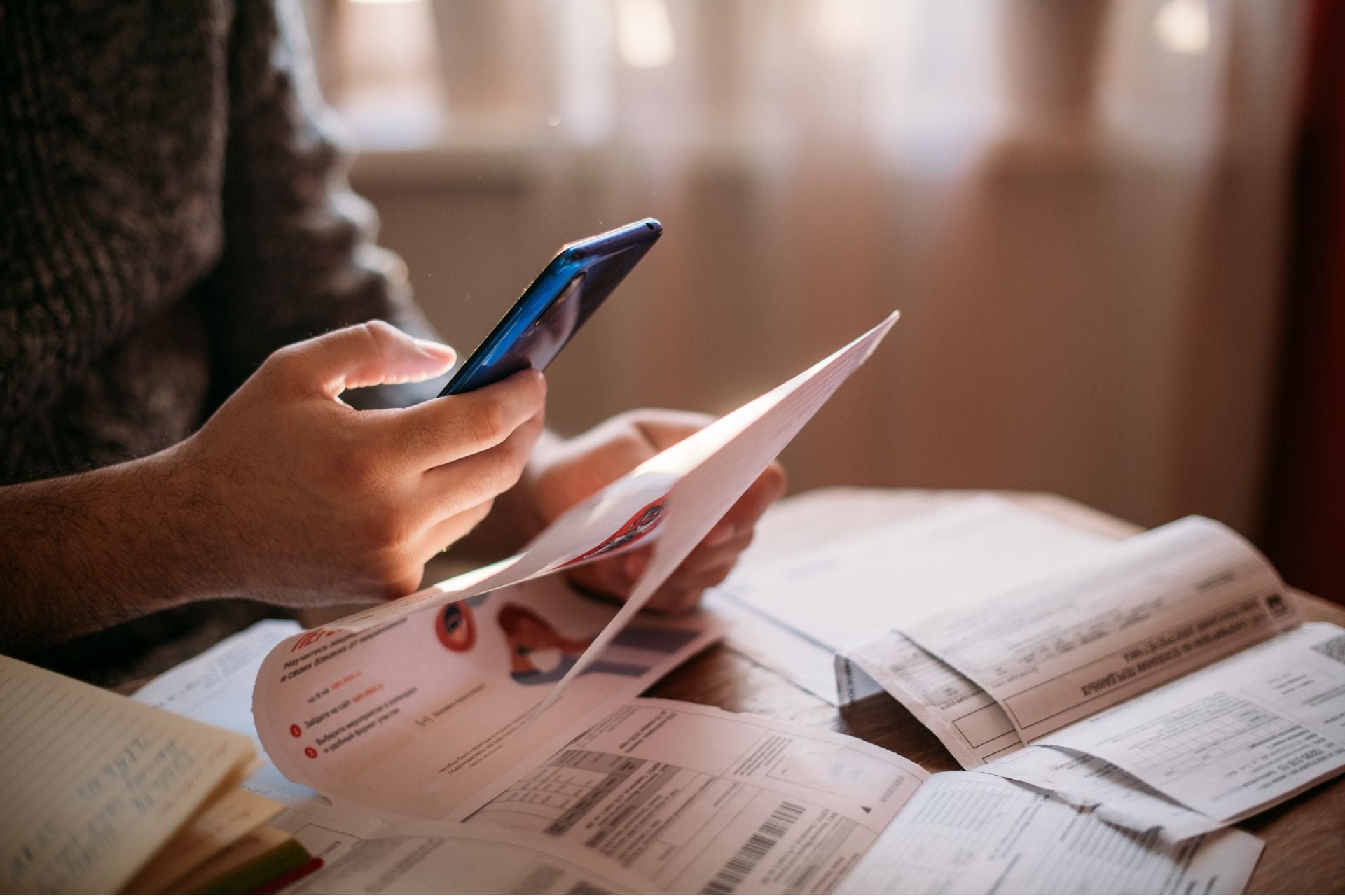 A person holding a smartphone and a bill with more bills spread out on the table