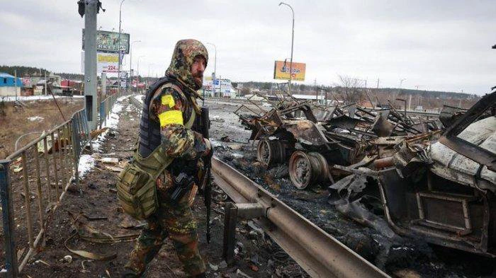 TERKINI Perang Rusia-Ukraina: Seorang pria bersenjata yang di lengannya ada tanda kain kuning berdiri di dekat sisa-sisa kendaraan militer Rusia di Bucha, dekat ibu kota Kyiv, Ukraina, Selasa (1/4/2022).