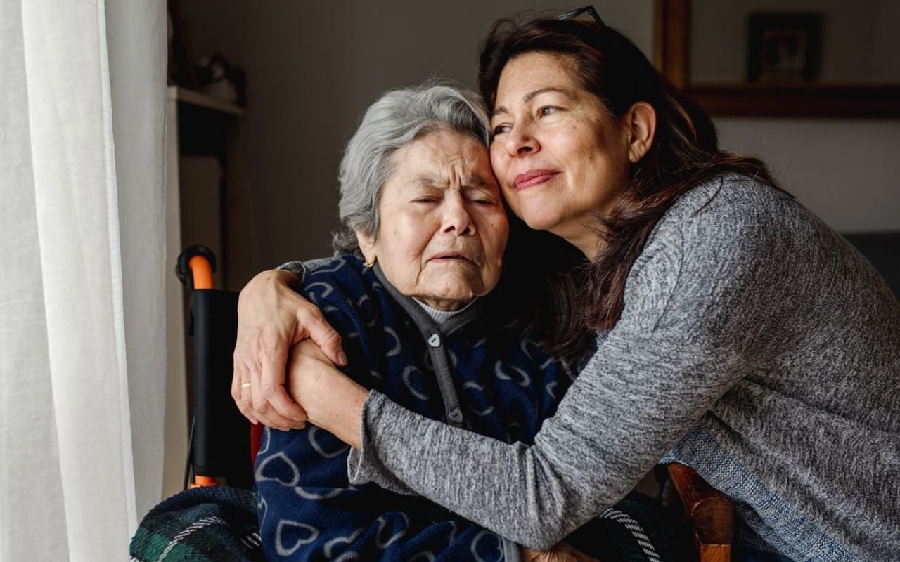 A daughter hugs her elderly mother who is in a wheelchair.