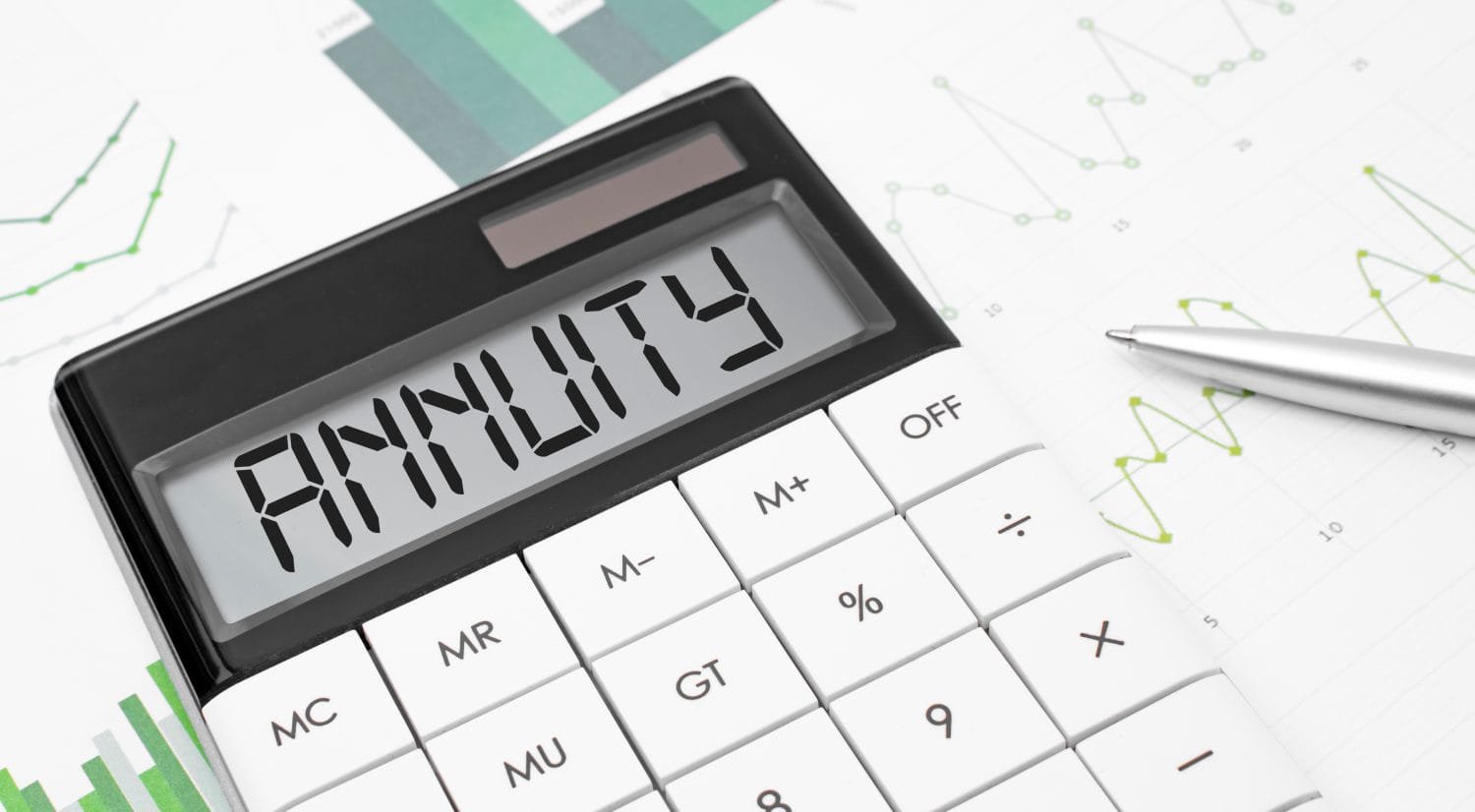 A close-up view of a black calculator with white keys. The calculator's digital screen displays the word 'ANNUITY' in large black capital letters. The calculator is placed on a white document featuring green line graphs and green bar charts. A silver pen rests on the document to the right of the calculator.