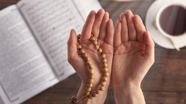 female hand of prayer with wooden beads in sunlight, iftar concept, Ramadan month, Koran, plate of dried fruit, Cup of tea on wooden table