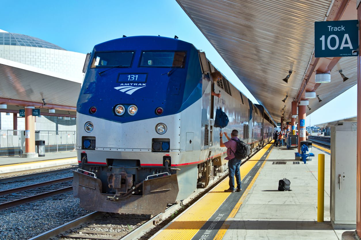 A passenger hands a bag to a crew member from the door of an Amtrak train at a station platform labeled Track 10A.