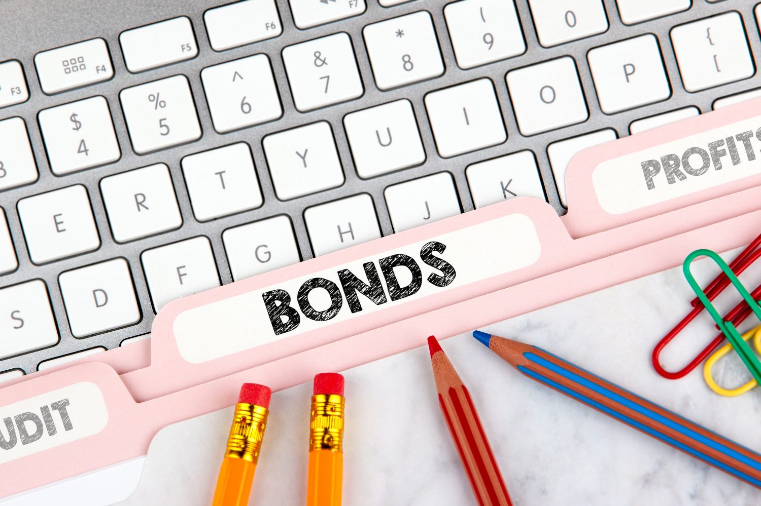 An overhead shot of a laptop keyboard and pink file folders. The central file tab prominently displays 'BONDS' in bold black letters, while partially visible tabs to the left and right read 'AUDIT' and 'PROFITS' respectively. Two yellow pencils with red erasers and colorful paper clips rest on a white marble-patterned surface in the foreground.