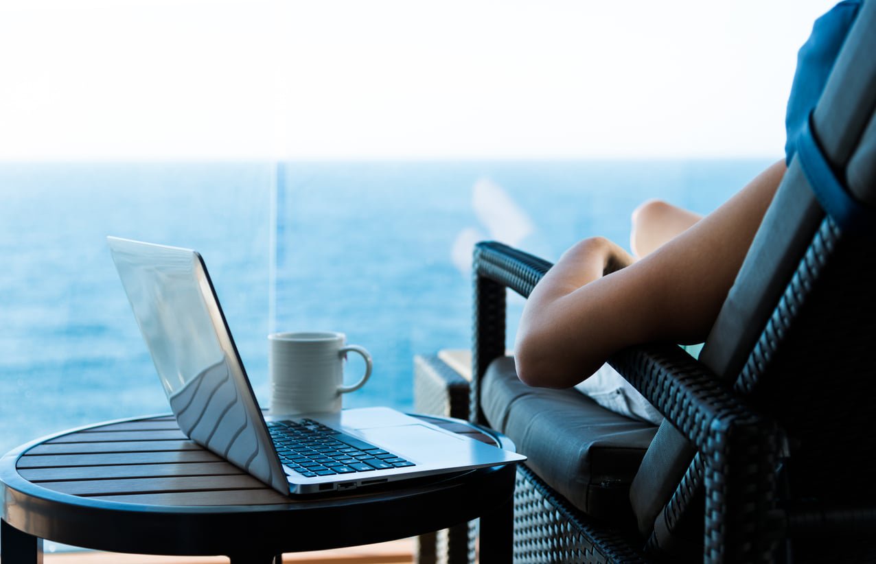 A person sitting in a chair on a balcony with a laptop and a mug on a small table, overlooking the ocean.