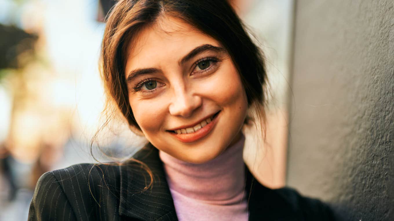 Portrait of a frugal brunette woman wearing a casual blazer, leaning on a wall outside. 
