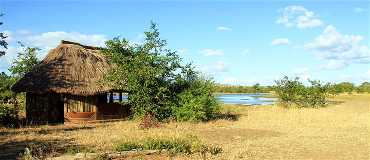 A thatched-roof, open-sided accommodation structure on the banks of a river; some trees shade the building.