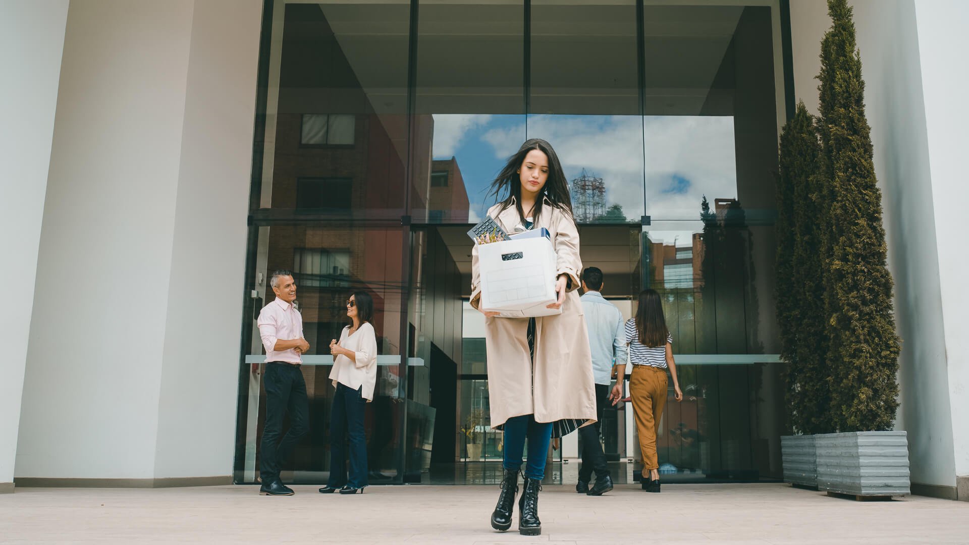 Discover a fired woman leaving the office building with her belongings in a box and looking sad