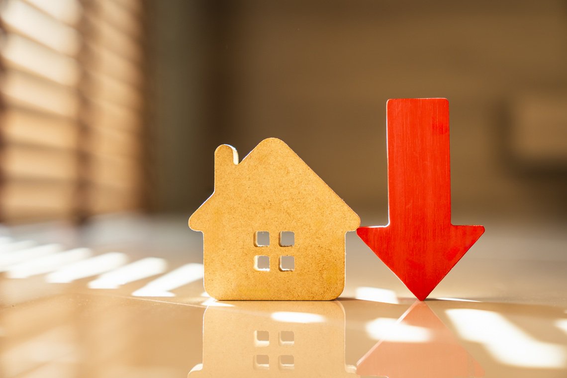 Real estate housing market decline concept. Wooden house model standing next to falling red arrow on floor with window blind shadows. Decreasing property value and mortgage rate.