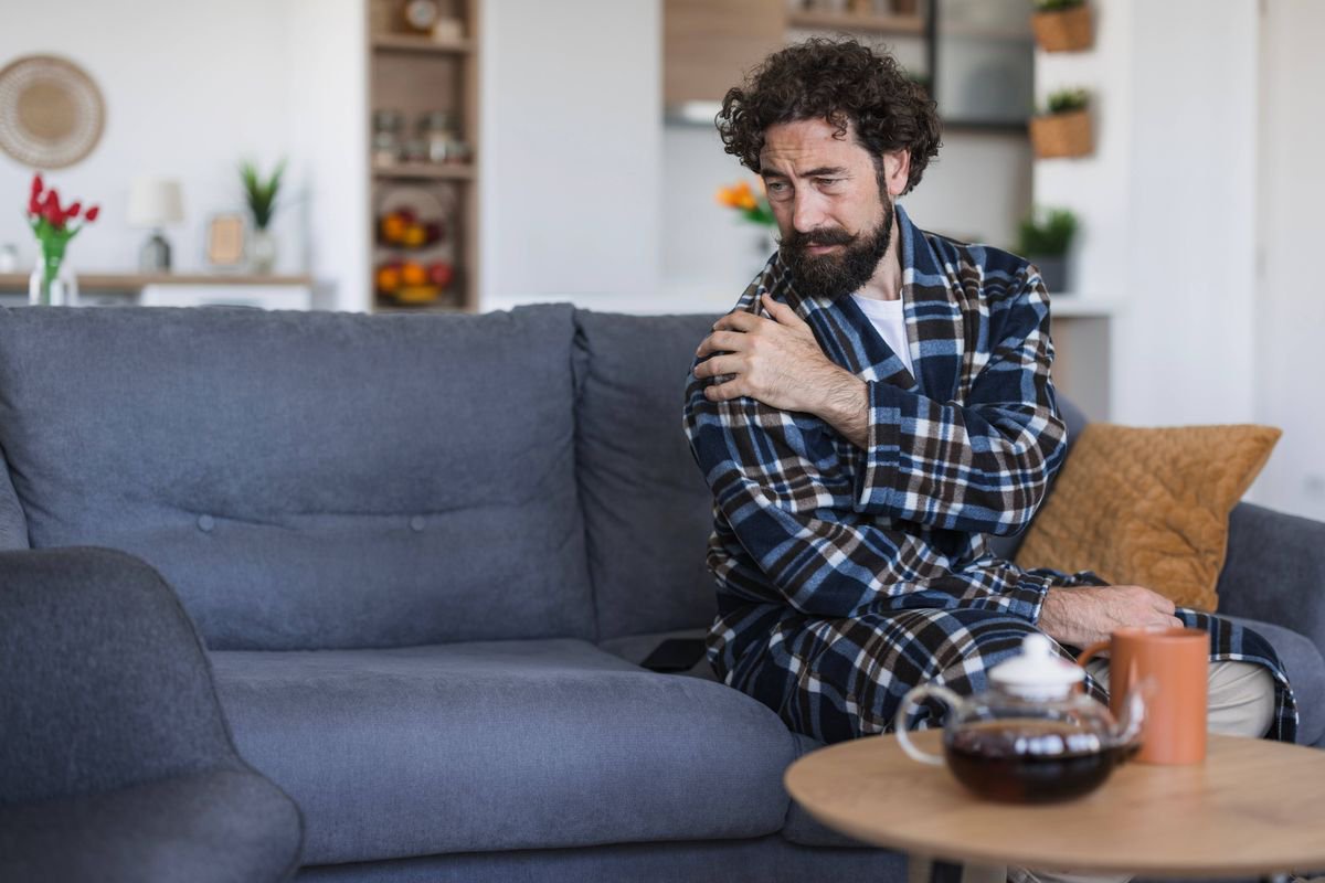 Man in bathrobe experiencing discomfort, touching his shoulder while sitting on couch