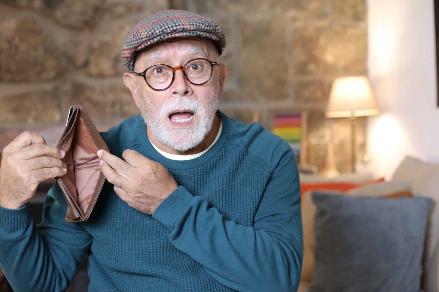 A senior man with a white beard, round glasses, and a plaid newsboy cap holds an open, empty brown leather wallet in both hands, looking at the viewer with wide eyes and an open mouth in a shocked expression. He is wearing a teal blue knitted sweater. The blurred background suggests an indoor setting with a stone wall and a lit lamp.