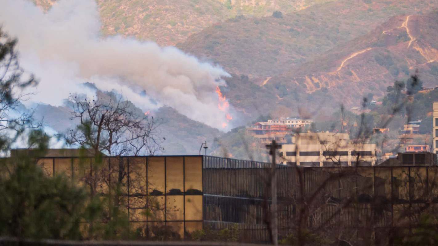 Smoke and flames rise from a hillside with buildings visible in the foreground.