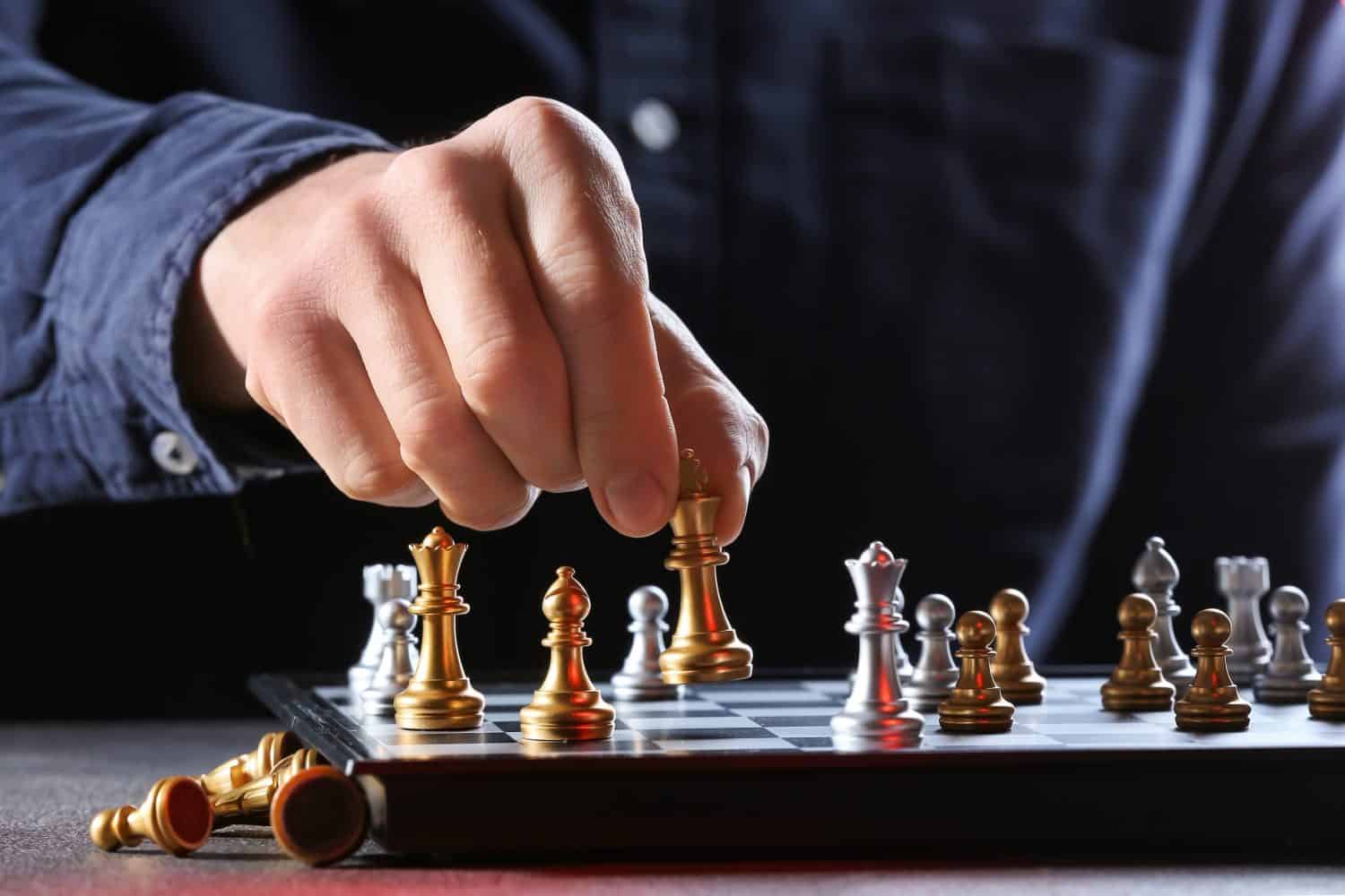 A person's hand, wearing a blue long-sleeved shirt, is holding a golden chess king piece, poised to place it on a chessboard. Other gold and silver chess pieces are arranged on the board, some in sharp focus while others are softly blurred in the foreground and background.