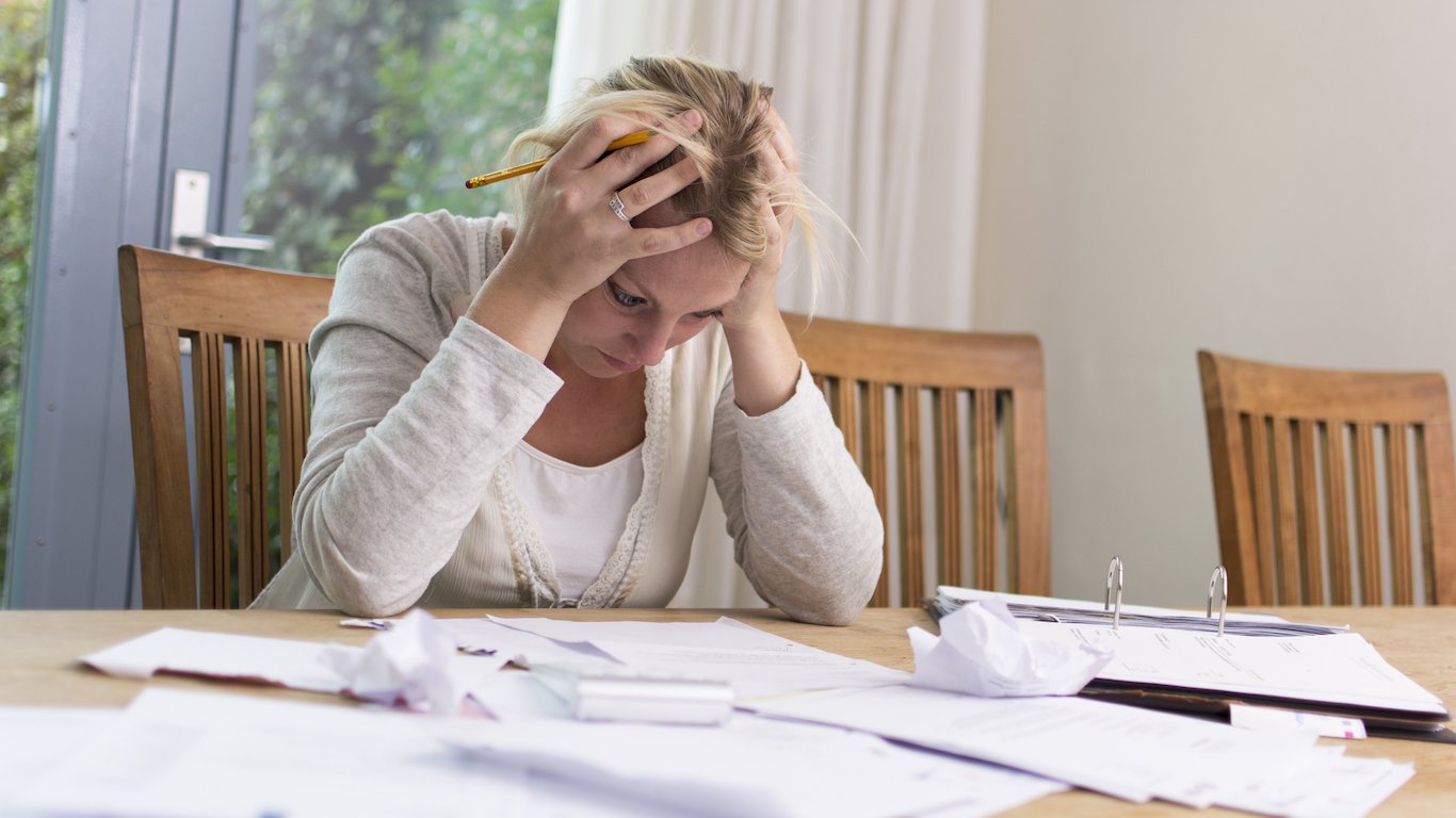 A blonde woman in a light beige cardigan sits at a wooden table, head bowed, with her hands clasped on top of her head in a gesture of despair or stress. A yellow pencil is tucked into her hair. Numerous papers, some crumpled, and an open binder are scattered across the table in front of her. The background features a large window with greenery outside on the left and a plain light-colored wall on the right.