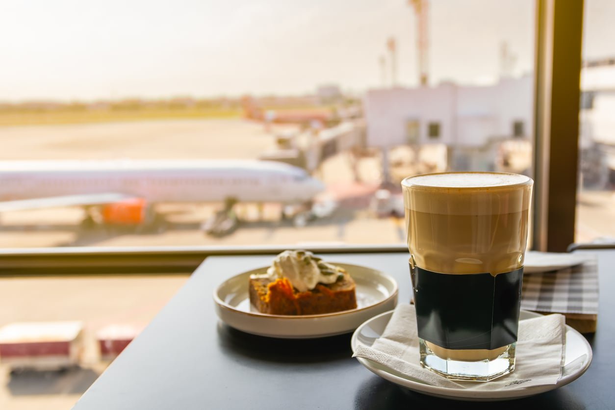 A glass of latte and a small cake with cream on a table overlooking an airport tarmac with an airplane.