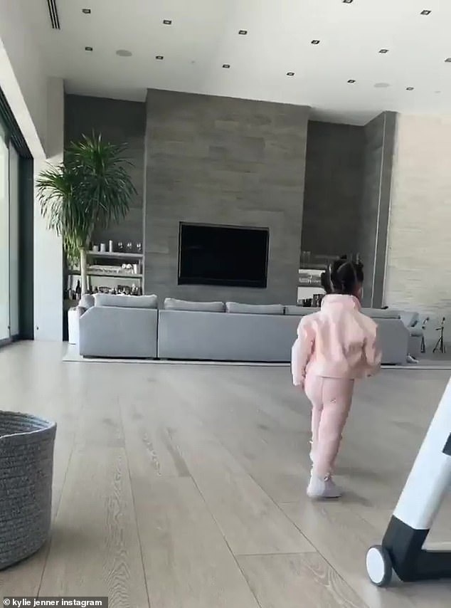 ABCDEFG Bye! The little girl does not stop to pose for her mother but rather runs off to the living room which is all grey with a large TV on the wall and recessed lighting