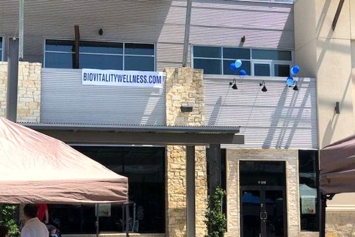 Building exterior with "BIOVITALITYWELLNESS.COM" banner, blue balloons, stone pillars, and tents below.